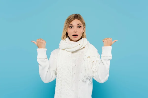 Confused blonde woman in white sweater and scarf pointing with thumbs isolated on blue — Stock Photo