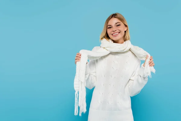Happy blonde woman in white sweater adjusting winter scarf isolated on blue — Stock Photo