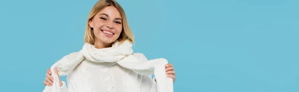 Щаслива блондинка в білому светрі, що регулює зимовий шарф ізольована на синьому, банер — стокове фото