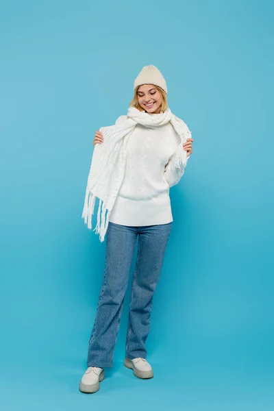 Pleine longueur de femme blonde joyeuse en pull blanc et chapeau ajustant écharpe d'hiver sur bleu — Photo de stock