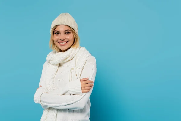 Счастливая блондинка в белом свитере и зимней шляпе, стоящая со скрещенными руками на голубом фоне — стоковое фото