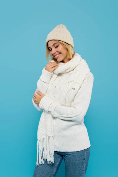 Mujer rubia feliz en suéter blanco y sombrero de invierno sonriendo aislado en azul - foto de stock