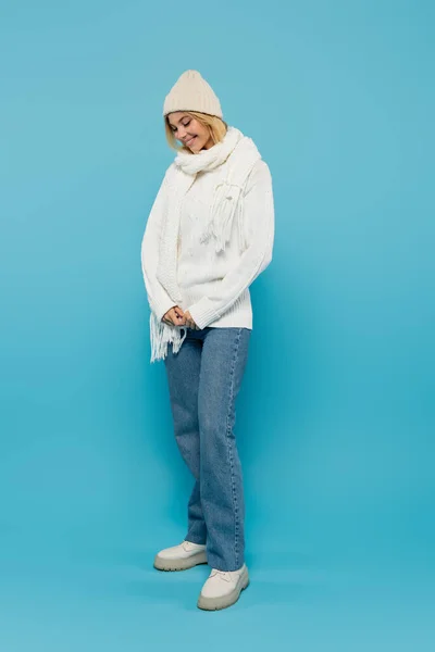 Pleine longueur de femme blonde timide en pull blanc et chapeau d'hiver souriant sur bleu — Photo de stock