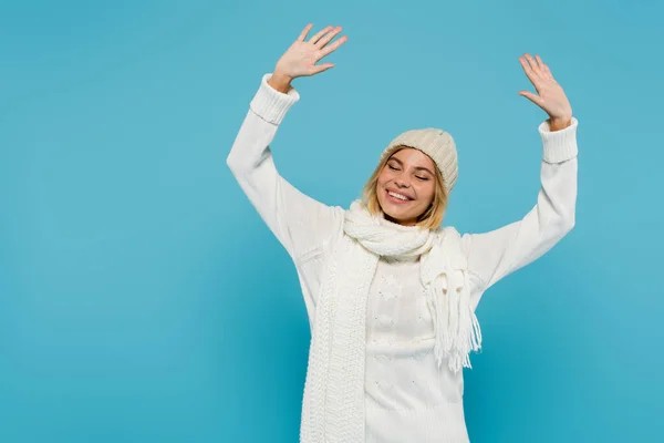 Mujer rubia positiva en suéter blanco y sombrero de invierno levantando las manos aisladas en azul - foto de stock