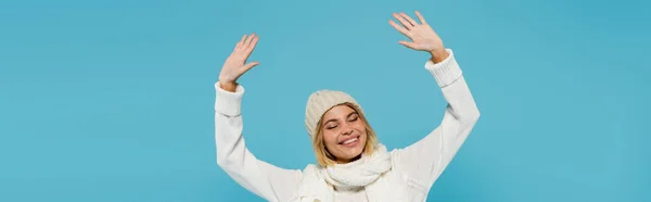 Positive blonde woman in white sweater and winter hat raising hands isolated on blue, banner — Stock Photo