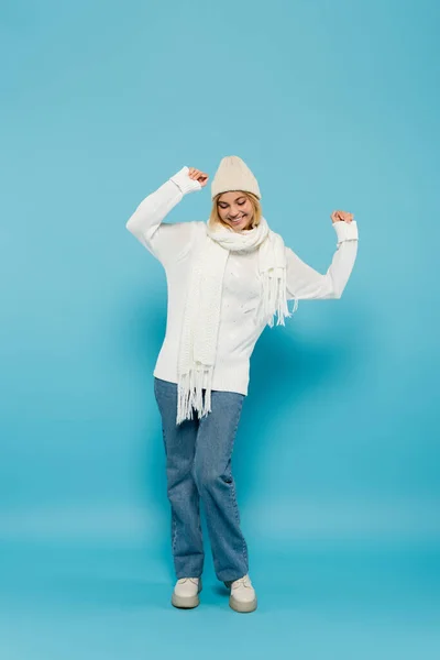 Pleine longueur de femme blonde positive en pull blanc et chapeau d'hiver dansant sur bleu — Photo de stock