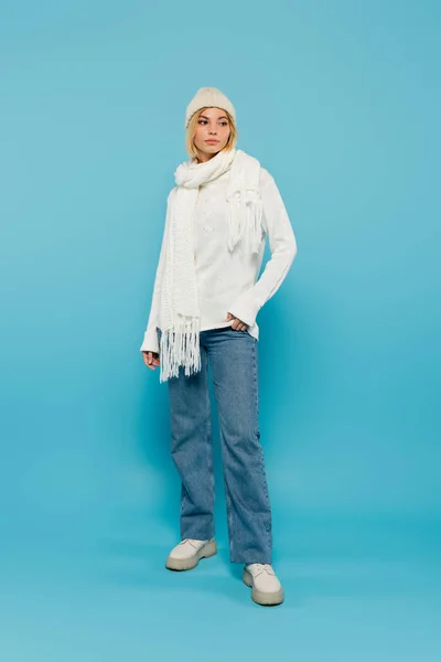 Longitud completa de mujer rubia joven en suéter blanco y sombrero de invierno de pie con la mano en el bolsillo en azul - foto de stock