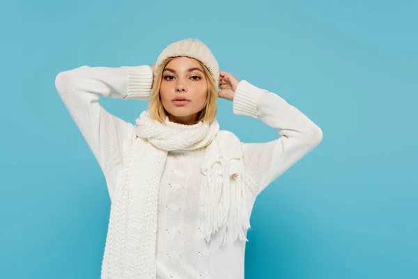 Young blonde woman in white sweater and winter scarf adjusting knitted hat isolated on blue — Stock Photo