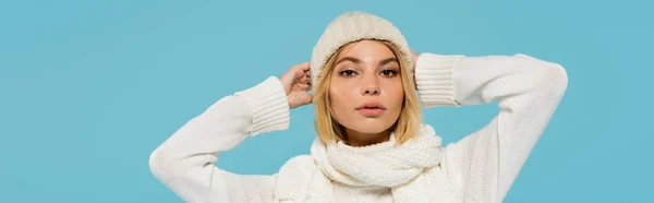 Young blonde woman in white sweater and winter scarf adjusting knitted hat isolated on blue, banner — Stock Photo