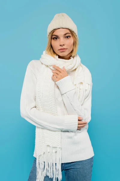 Portrait of young blonde woman in white sweater, scarf and knitted hat looking at camera isolated on blue — Stock Photo