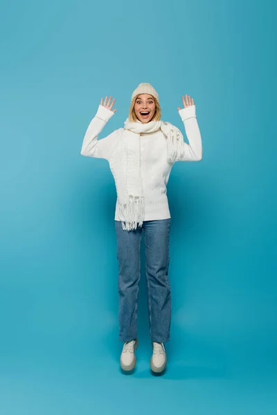 Full length of excited young woman in winter outfit and opened mouth levitating on blue — Stock Photo