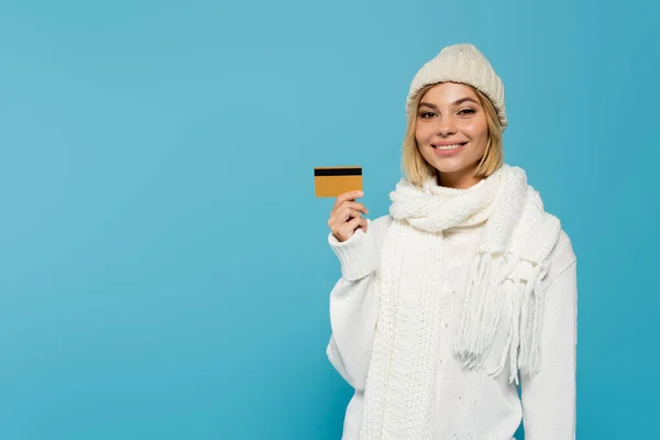 Happy blonde woman in white sweater and winter hat holding credit card isolated on blue — Stock Photo