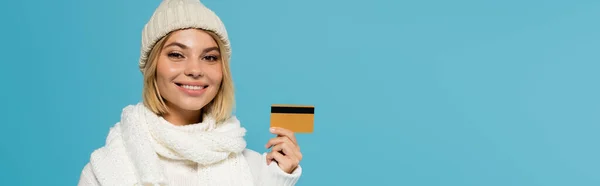 Mujer rubia feliz en suéter blanco y sombrero de invierno con tarjeta de crédito aislada en azul, pancarta - foto de stock