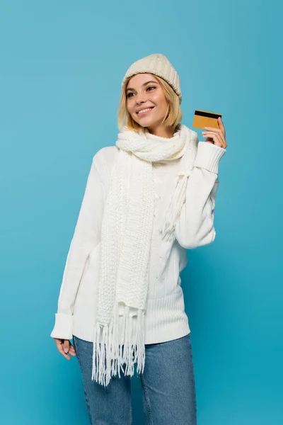 Cheerful young woman in white sweater and winter hat holding credit card isolated on blue — Stock Photo