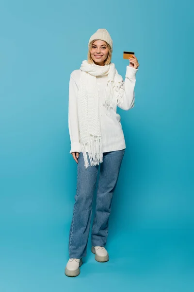 Pleine longueur de femme blonde heureuse en pull blanc et chapeau d'hiver tenant carte de crédit sur bleu — Photo de stock