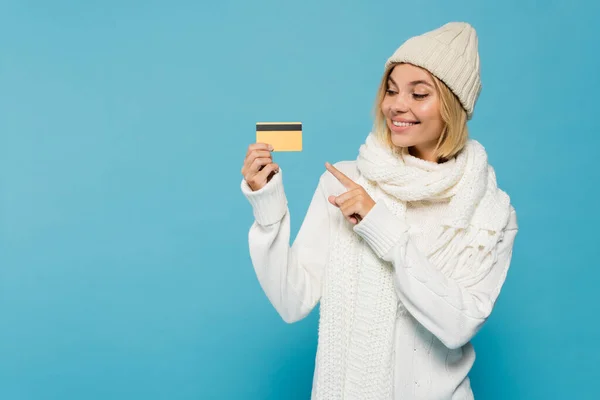 Happy blonde woman in white sweater and winter hat pointing with finger at credit card isolated on blue — Stock Photo