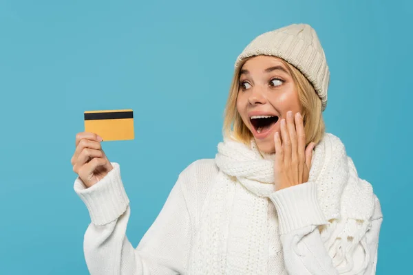 Mulher animado em suéter branco e chapéu de inverno segurando cartão de crédito isolado em azul — Fotografia de Stock