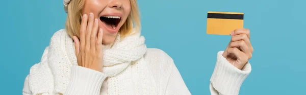 Обрезанный вид изумленной женщины в белом свитере и зимнем шарфе, держащей кредитную карту изолированы на синий, баннер — стоковое фото
