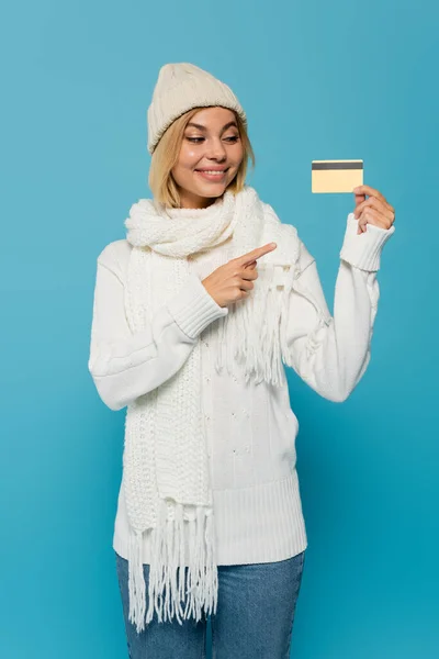 Positive woman in white sweater and winter hat pointing with finger at credit card isolated on blue — Stock Photo