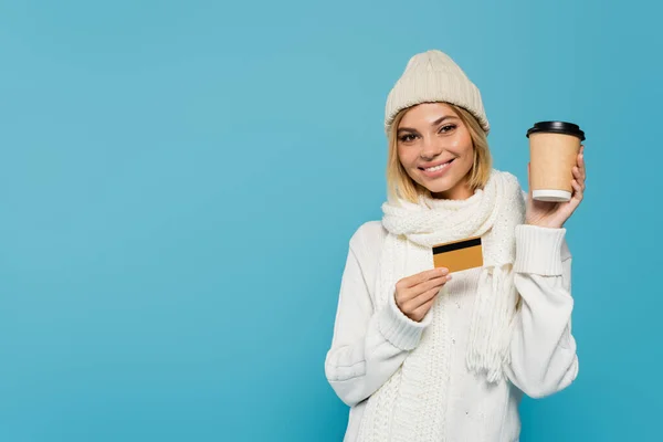 Happy blonde woman in white winter outfit holding credit card and paper cup isolated on blue — Stock Photo