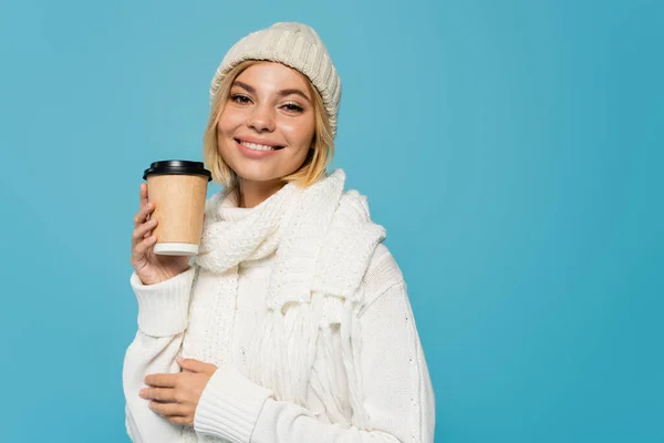 Cheerful woman in white sweater and knitted hat holding paper cup with takeaway drink isolated on blue — Stock Photo