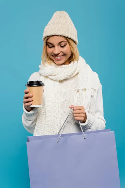 Щаслива блондинка в білому светрі і в'язаний капелюх тримає паперову чашку і сумку ізольовані на синьому — стокове фото