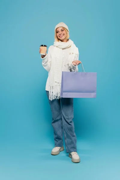 Longitud completa de mujer rubia feliz en suéter blanco y sombrero de punto sosteniendo taza de papel y bolsa de compras en azul - foto de stock