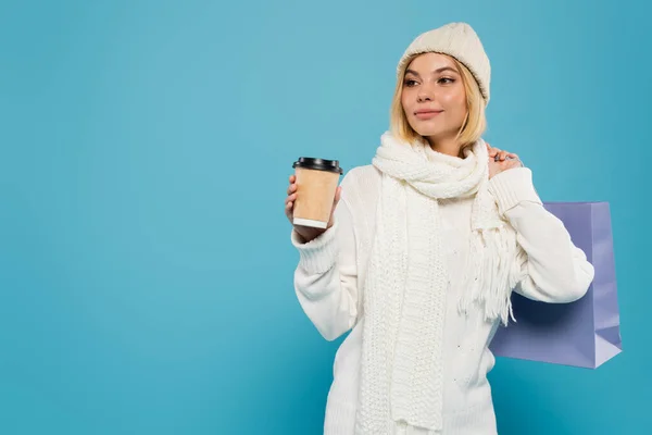 Jovem mulher em suéter branco e chapéu de malha segurando copo de papel e saco de compras isolado em azul — Fotografia de Stock