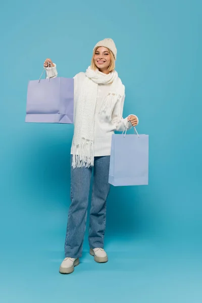 Pleine longueur de femme gaie en pull blanc et chapeau tricoté tenant des sacs à provisions sur bleu — Photo de stock