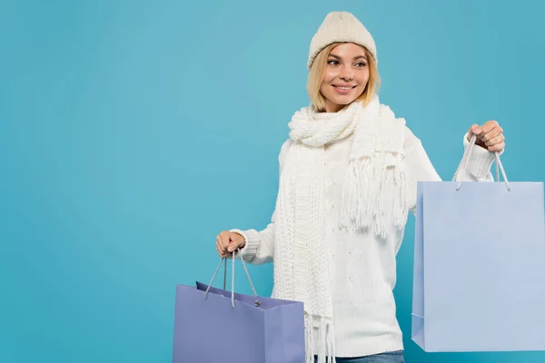 Jovem alegre em camisola branca e chapéu de malha segurando sacos de compras isolados em azul — Fotografia de Stock