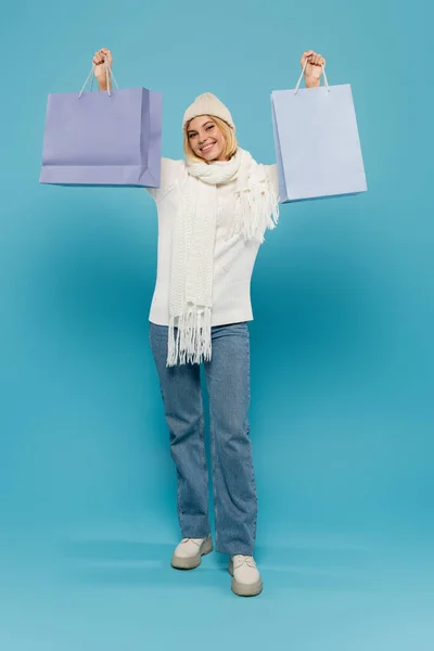 Pleine longueur de joyeuse jeune femme en pull blanc et chapeau tricoté tenant des sacs à provisions sur bleu — Photo de stock