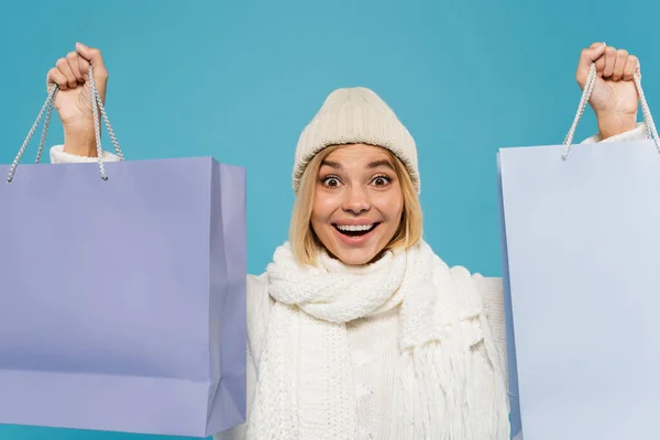 Mujer joven excitada en suéter blanco y sombrero de punto con bolsas de compras aisladas en azul — Stock Photo