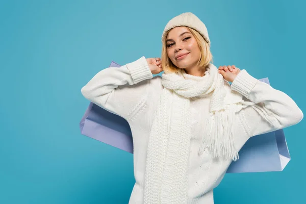 Smiling young woman in white sweater and knitted hat holding shopping bags isolated on blue — Stock Photo