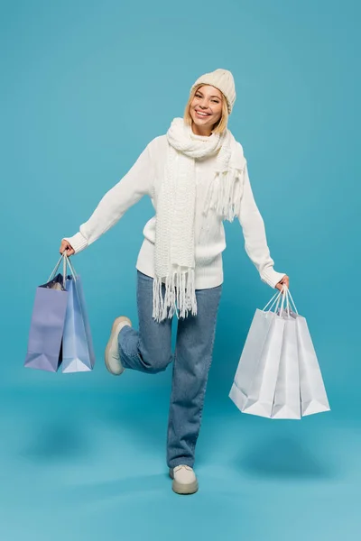 Pleine longueur de jeune femme positive en pull blanc et chapeau tenant des sacs à provisions sur bleu — Photo de stock