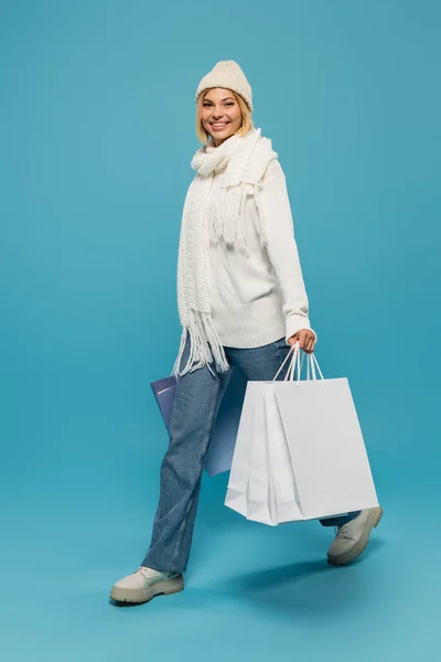Полная длина радостной молодой женщины в белом свитере и шляпе держа сумки для покупок на синий — стоковое фото