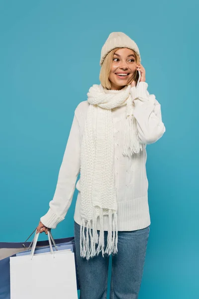 Gioiosa giovane donna in maglione bianco e cappello che tiene borse della spesa mentre parla su smartphone isolato su blu — Foto stock
