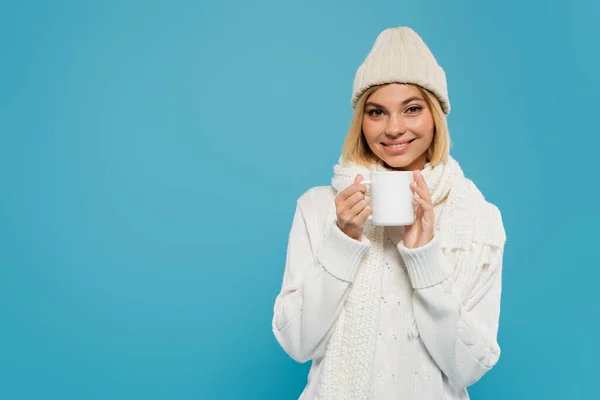 Mulher alegre em suéter branco e chapéu segurando xícara de café isolado em azul — Fotografia de Stock