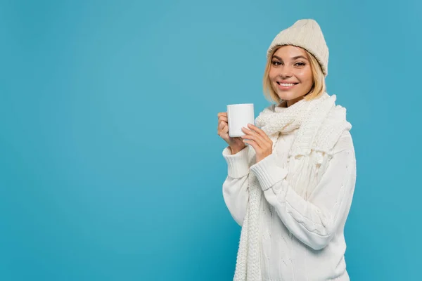 Alegre joven en suéter blanco y sombrero sosteniendo taza de café aislado en azul - foto de stock