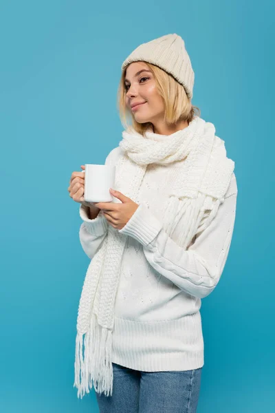 Smiling woman in white sweater and winter hat holding cup of coffee isolated on blue — Stock Photo