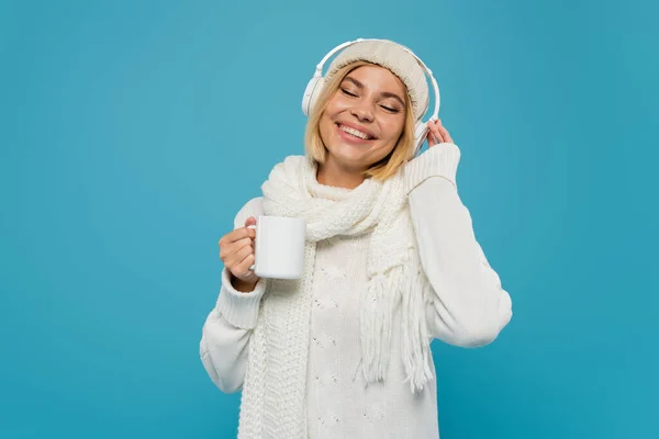Femme souriante en pull blanc et chapeau tenant une tasse de café tout en écoutant de la musique dans des écouteurs sans fil isolés sur bleu — Photo de stock