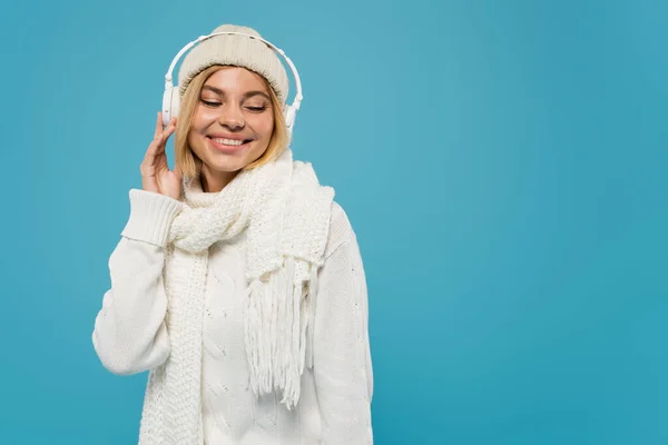 Lächelnde Frau in weißem Pullover und Hut, die Musik in drahtlosen Kopfhörern isoliert auf blauem Grund hört — Stockfoto