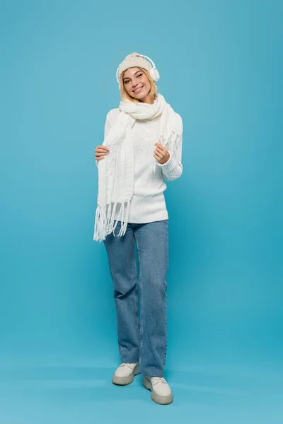 Comprimento total da mulher sorridente em suéter branco e chapéu ouvindo música em fones de ouvido sem fio em azul — Fotografia de Stock