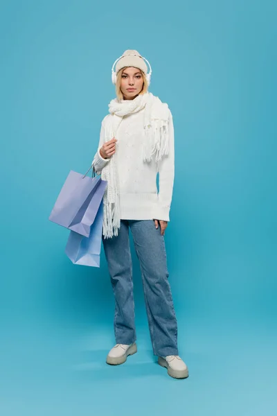 Full length of young woman in winter outfit and wireless headphones holding shopping bags on blue — Stock Photo