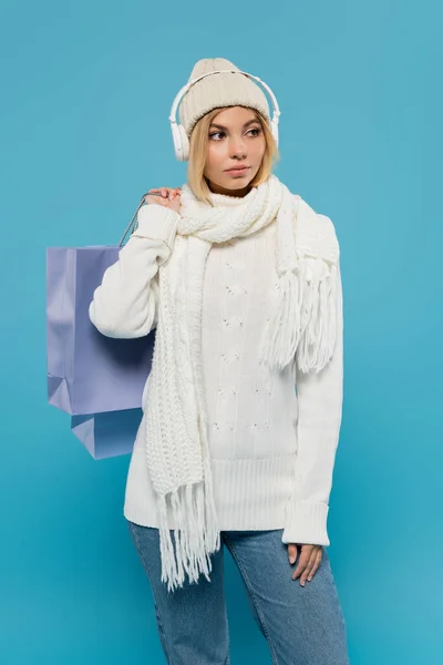 Blonde woman in winter outfit and wireless headphones holding shopping bags isolated on blue — Stock Photo
