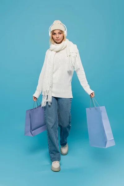 Full length of blonde woman in winter outfit and wireless headphones walking with shopping bags on blue — Stock Photo