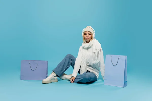 Blonde Frau in winterlichem Outfit und kabellosen Kopfhörern in der Nähe von Einkaufstaschen auf blauem Grund — Stockfoto