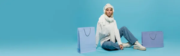 Blonde Frau in winterlichem Outfit und kabellosen Kopfhörern in der Nähe von Einkaufstaschen auf blauem Grund, Banner — Stockfoto
