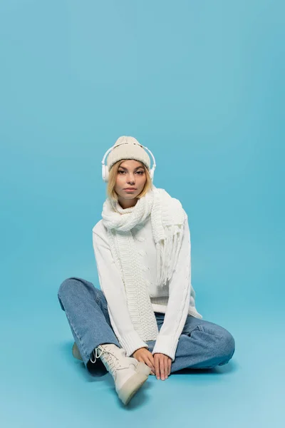 Full length of blonde woman in winter outfit and wireless headphones sitting on blue — Stock Photo