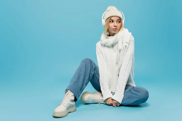 Full length of stylish blonde woman in winter outfit and wireless headphones sitting on blue — Stock Photo