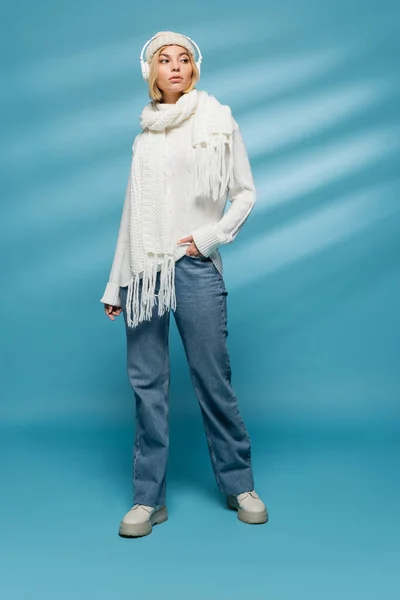 Полная длина стильная молодая женщина в зимнем наряде и беспроводные наушники позируют с рукой в кармане на синий — стоковое фото